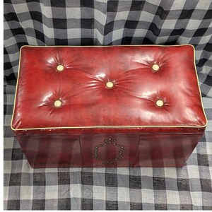 Puede incluir: Un otom&aacute;n rectangular rojo con una parte superior acolchada y botones dorados. Los bordes est&aacute;n adornados con un borde dorado. La parte delantera del otom&aacute;n presenta un dise&ntilde;o decorativo de peque&ntilde;os tachuelas redondas.