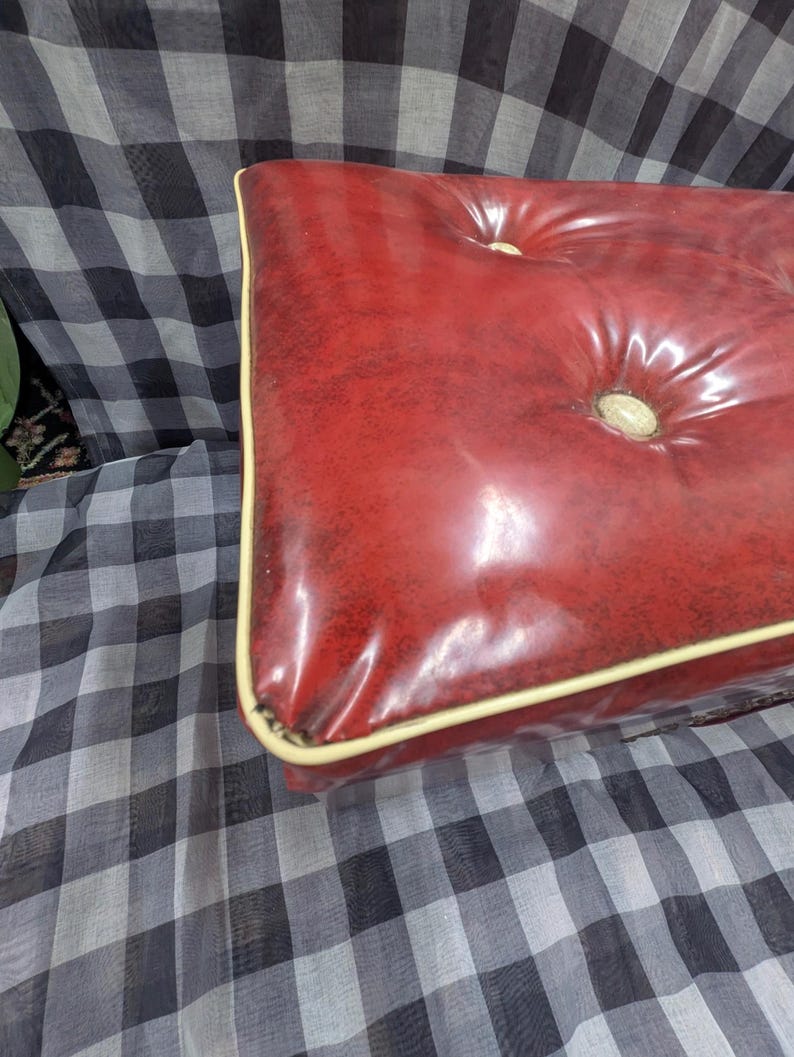 Puede incluir: Un otom&aacute;n rectangular, rojo y acolchado con un acabado brillante. El otom&aacute;n tiene un ribete color crema y detalles de botones. El fondo es un patr&oacute;n de cuadros blanco y negro.