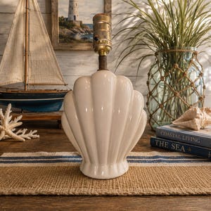 May include: A white ceramic table lamp base shaped like a large scallop shell. The lamp features a vertical ribbed design with a glossy finish and a metallic brass-colored socket assembly extending from the top center of the shell structure.