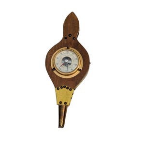 May include: A vintage wooden barometer with a brass-colored bezel and a gold-colored fabric accent. The barometer has a dark wood finish and a circular gauge with numbers and a needle. The handle is a dark wood color.