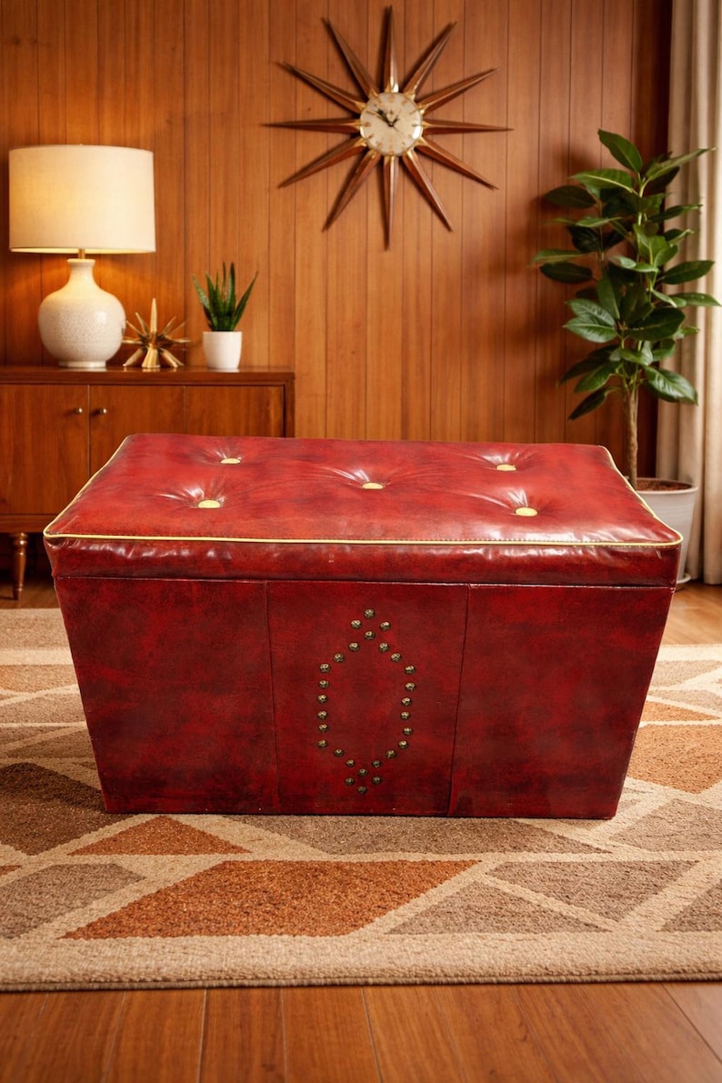 Puede incluir: Un otom&aacute;n de cuero rojo con ribete dorado y capiton&eacute;. La parte delantera del otom&aacute;n presenta un dise&ntilde;o decorativo con tachuelas. El otom&aacute;n est&aacute; colocado sobre una alfombra estampada. Una pared de madera y un reloj de estrella en el fondo.