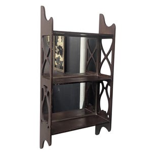 May include: A dark brown, three-tiered wooden shelf with decorative cutouts and a mirrored backing. The shelf has a rectangular shape with two shelves and a top section with a curved design. The shelf is designed to stand upright.