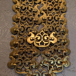 May include: A collection of ornate, brass-colored drawer pulls with intricate scrollwork and decorative handles. The hardware is arranged in a pattern, showcasing the detailed design and vintage aesthetic. The pulls are likely for furniture restoration or decorative projects.