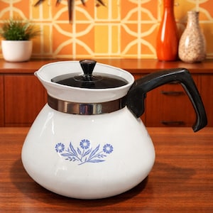 May include: White vintage teapot with a black handle and lid. The teapot has a floral design in blue and a silver band around the middle. The teapot is sitting on a wooden surface.