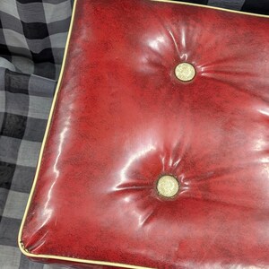 Puede incluir: Un otom&aacute;n rojo acolchado con ribete color crema. El coj&iacute;n rectangular tiene dos botones y un acabado brillante. El otom&aacute;n est&aacute; sobre un fondo a cuadros blanco y negro.