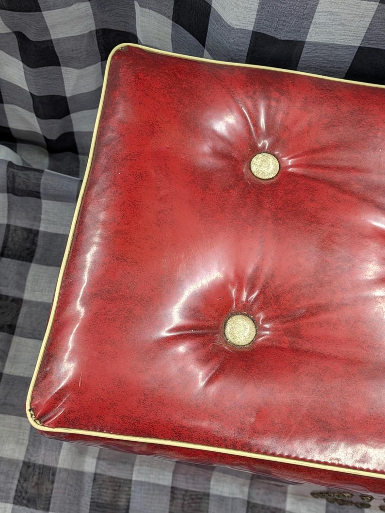 Puede incluir: Un otom&aacute;n rojo acolchado con ribete color crema. El coj&iacute;n rectangular tiene dos botones y un acabado brillante. El otom&aacute;n est&aacute; sobre un fondo a cuadros blanco y negro.