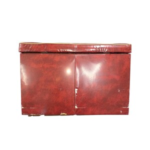 Puede incluir: Un banco de almacenamiento rectangular vintage con un exterior rojo oscuro texturizado. El borde superior tiene un fino ribete dorado. El banco parece estar hecho de un material duradero, con algo de desgaste visible en los bordes.
