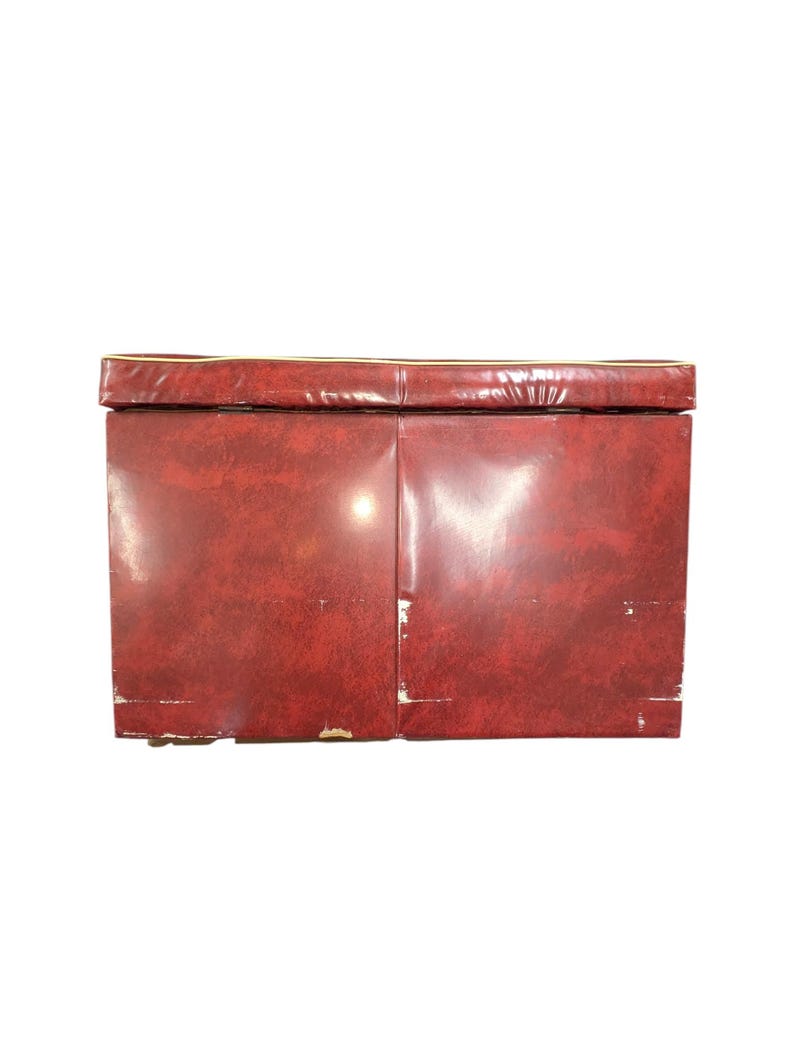 Puede incluir: Un banco de almacenamiento rectangular vintage con un exterior rojo oscuro texturizado. El borde superior tiene un fino ribete dorado. El banco parece estar hecho de un material duradero, con algo de desgaste visible en los bordes.