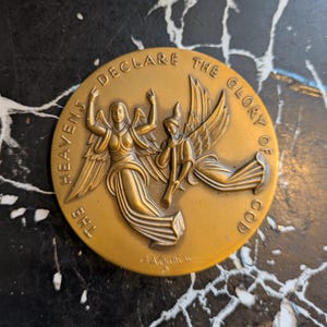 May include: A bronze-colored round medallion with the text "THE HEAVENS DECLARE THE GLORY OF GOD." The medallion features two winged figures, one with arms raised and the other playing a horn. The artist's signature is at the bottom.