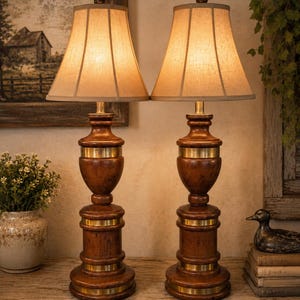 May include: A pair of table lamps featuring turned wooden bases with a warm brown finish and horizontal brass metal bands. Each lamp is topped with a flared, off-white fabric lampshade that emits a soft, warm glow from the internal light source.