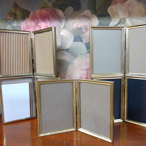 May include: A set of eight gold-toned picture frames with a decorative border. The frames are arranged in a row on a wooden surface. Some of the frames are open and show the cardboard backing.