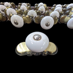 May include: A collection of white ceramic cabinet knobs with brass-colored bases. Each knob has a rounded, oval shape with a silver-colored center. The brass bases have a decorative, scalloped design.