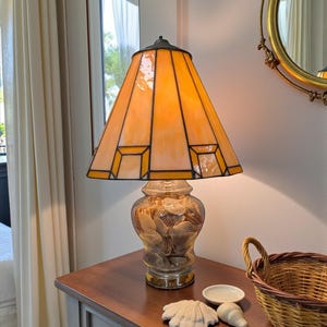 May include: A decorative table lamp with a stained glass shade in shades of orange and yellow. The base is a glass jar filled with seashells. The lamp sits on a wooden table with a small basket and decorative shells.