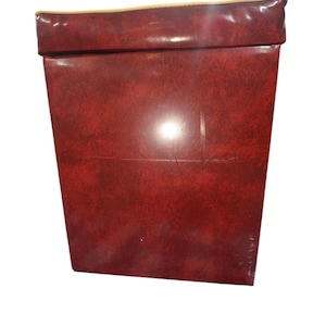 Puede incluir: Una caja rectangular roja con una superficie texturizada. El borde superior est&aacute; doblado y tiene un ribete dorado. La caja est&aacute; hecha de un material que parece ser cuero o similar al cuero.