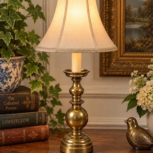 May include: A brass table lamp with a polished metallic finish, featuring a tiered, rounded base and a central stem. The lamp is topped with a cream-colored, bell-shaped fabric shade that has a textured surface and a decorative braided trim.