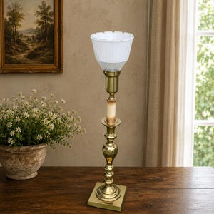 May include: A tall brass candlestick lamp featuring a square base, a slender turned stem, and a central faux candle sleeve. The top is fitted with a white, fluted glass shade that has a scalloped rim and a textured, cross-hatched pattern.