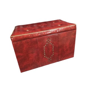 Puede incluir: Un otom&aacute;n de almacenamiento rojo con la parte superior acolchada y ribete dorado. El panel frontal presenta un dise&ntilde;o ovalado decorativo hecho de peque&ntilde;os tachuelas redondas. El otom&aacute;n tiene forma rectangular.