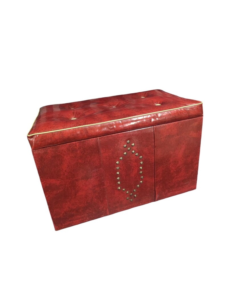 Puede incluir: Un otom&aacute;n de almacenamiento rojo con la parte superior acolchada y ribete dorado. El panel frontal presenta un dise&ntilde;o ovalado decorativo hecho de peque&ntilde;os tachuelas redondas. El otom&aacute;n tiene forma rectangular.