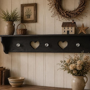 May include: A black wooden wall shelf featuring three clear glass knobs and two heart-shaped cutouts. The shelf has a flat top surface and curved side supports, providing a functional storage area for hanging items while maintaining a simple design.