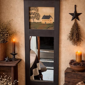 May include: A rectangular black framed mirror with a painted landscape scene at the top. The painting depicts a yellow house with a black roof, a tree, and green grass. The mirror reflects a room with a table and chair. The style is rustic and country.