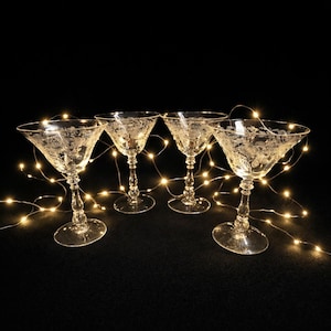 May include: Four clear, etched glass martini glasses with delicate floral patterns. The glasses are arranged on a dark surface, illuminated by warm, glowing fairy lights. The stemware has a classic, elegant design.