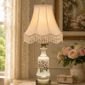 May include: A table lamp with a white ceramic base featuring a pink rose floral pattern and gold trim. The lamp has a beige fabric shade with a scalloped edge, clear crystal fringe, and a decorative gold metal finial at the top of the shade.