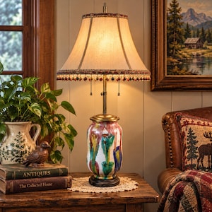 May include: A table lamp with a brass base and a ceramic body featuring vertical drips of green, blue, and red paint on a white background. The lamp has a beige fabric shade with a decorative beaded fringe trim and two pull-chain switches.