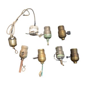 May include: A collection of eight vintage brass light bulb sockets with various styles and finishes. Some have a green patina, while others are polished. The sockets are in various states of wear and tear, adding to their vintage charm.