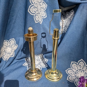 May include: Two gold candle holders with a simple design. The candle holders are on a blue fabric background with white floral embroidery.