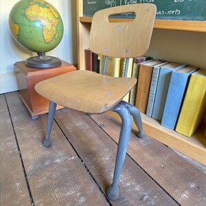 May include: A vintage wooden school chair with a light-colored seat and back, and gray metal legs. The chair is positioned on a wooden floor. A globe and books are in the background.
