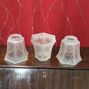 May include: Three frosted glass lamp shades with ornate floral designs. The shades have a hexagonal shape and are arranged on a dark wooden surface against a red wallpaper background.