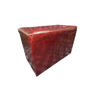 Puede incluir: Un otom&aacute;n de cuero burdeos con forma rectangular y parte superior acolchada. Los bordes est&aacute;n ribeteados con ribetes dorados y la parte delantera presenta tachuelas met&aacute;licas decorativas. Esta pieza de mobiliario es adecuada para una sala de estar o un dormitorio.