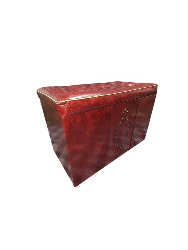 Puede incluir: Un otom&aacute;n de cuero burdeos con forma rectangular y parte superior acolchada. Los bordes est&aacute;n ribeteados con ribetes dorados y la parte delantera presenta tachuelas met&aacute;licas decorativas. Esta pieza de mobiliario es adecuada para una sala de estar o un dormitorio.