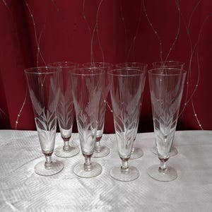 May include: A set of eight clear champagne flutes with etched leaf designs. The flutes have a tall, slender shape with a round base and a delicate stem. The background features a red curtain.