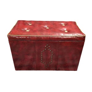Puede incluir: Un otom&aacute;n de cuero rojo con la parte superior acolchada y ribete dorado. Los lados presentan un patr&oacute;n decorativo de tachuelas met&aacute;licas. El otom&aacute;n es una unidad de almacenamiento y tiene forma rectangular.