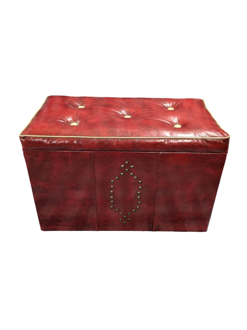 Puede incluir: Un otom&aacute;n de cuero rojo con la parte superior acolchada y ribete dorado. Los lados presentan un patr&oacute;n decorativo de tachuelas met&aacute;licas. El otom&aacute;n es una unidad de almacenamiento y tiene forma rectangular.