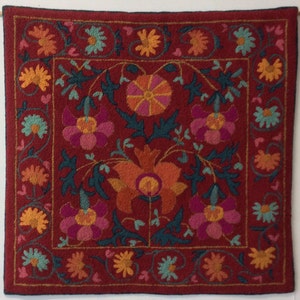 May include: A red wool tapestry with a floral design. The tapestry features a central flower with orange, pink, and yellow petals. The border of the tapestry is decorated with smaller flowers in shades of orange, yellow, and blue.