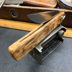 May include: A folding pocket knife with a light brown wooden handle and a silver blade. The knife is resting on a metal tool, with other woodworking tools visible in the background. The knife is a tool for cutting.