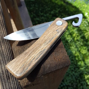 May include: A folding pocket knife with a wooden handle and a stainless steel blade. The knife has a hook on the end and is resting on a wooden surface. The blade is partially open, revealing a shiny, sharp edge. The handle has a natural wood grain pattern.