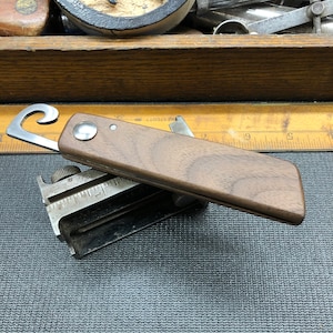 May include: A brown wooden handled pocket knife with a metal loop opener and a silver metal body. The knife is lying on a gray textured surface.