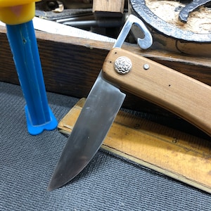 May include: A wooden-handled folding knife with a silver blade and a decorative silver emblem. The knife is partially open, revealing the sharp edge. A blue and yellow Pez dispenser is in the background.