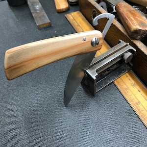 May include: A wooden handled straight razor with a silver blade, resting on a black surface with a ruler and a metal tool. The razor is partially open, revealing the blade.