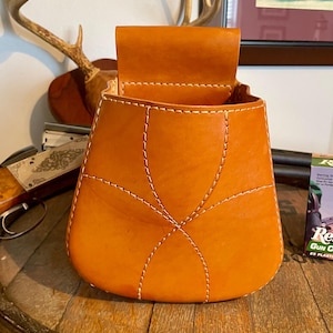 May include: An orange leather cartridge bag with white stitching. The bag has a flap closure and is open, revealing the interior. The bag is sitting on a wooden surface.