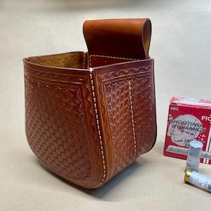 May include: A brown leather cartridge bag with a textured basketweave pattern and a flap closure. The bag is stitched with white thread and is next to a box of shotgun shells.