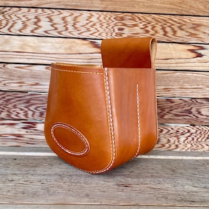 May include: A handcrafted, light brown leather pouch with white stitching. The pouch has a curved oval design on the front and a flap closure. The item is sitting on a wooden surface.
