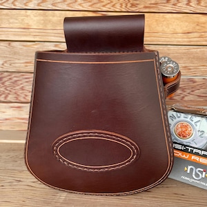 May include: A brown leather shotgun shell holder with a stitched oval design. The holder has a flap on the top and a strap on the side.
