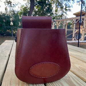 May include: A brown leather pouch with a flap closure and a stitched oval on the front. The pouch is sitting on a wooden surface.