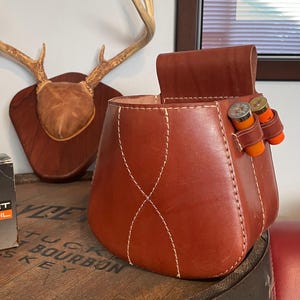 May include: A brown leather shotgun shell bag with white stitching and two orange and brass shotgun shells attached. The bag is sitting on a wooden barrel. A deer antler mount is in the background.
