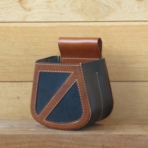 May include: A brown leather hanging pouch with a black leather flap and brown stitching. The pouch has a triangular shape with a curved bottom.