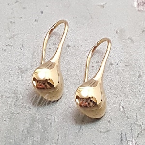 May include: A pair of gold-colored teardrop earrings. Each earring features a curved hook and a rounded, polished teardrop shape. The earrings are displayed against a textured, light gray background.